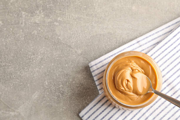 istock Glass jar with peanut butter and spoon, and kitchen towel on grey background, space for text and top view 1154356199