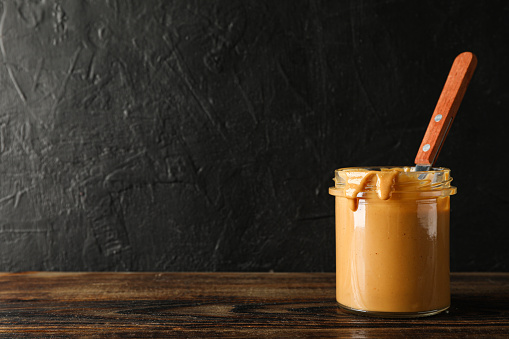 Glass jar with peanut butter and spoon on wooden table against black background, space for text