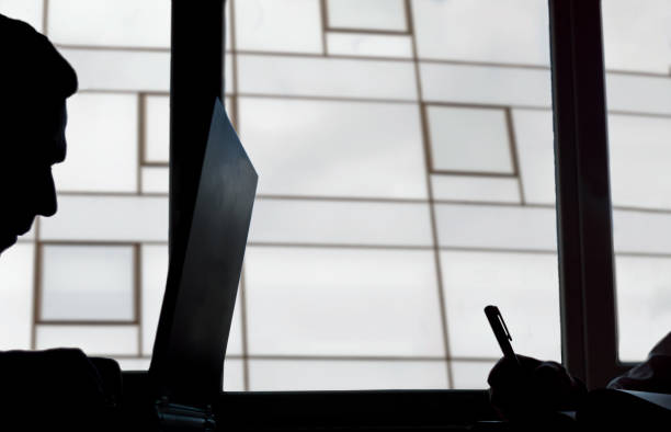 istock Silhouette of  a man keyboarding on laptop computer, while is sitting in modern office interior. 1154361306