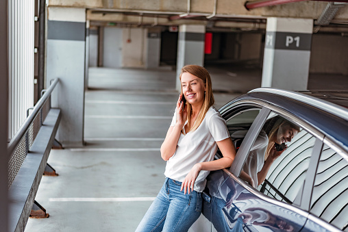 Photo of an attractive brown woman standing in the parking lot while talking on a smartphone near a new automobile. Smiling woman talking on a smartphone while standing near a new car. Portrait of a happy young woman standing while talking on the mobile phone.