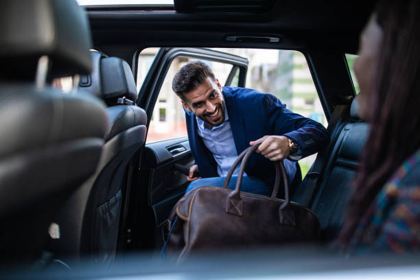 Good-looking man entering ride sharing car Young, handsome man entering a car, holding luggage, his friends sitting in car and waiting for him back seat photos stock pictures, royalty-free photos & images