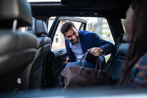 Young, handsome man entering a car, holding luggage, his friends sitting in car and waiting for him