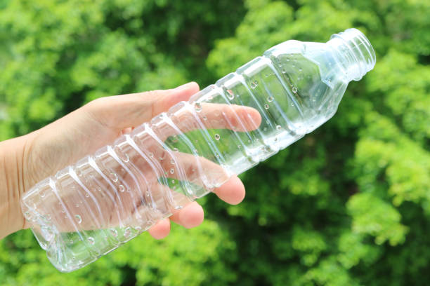 ręka człowieka trzymająca pustą plastikową butelkę wody pitnej przed rozmytymi zielonymi liśćmi - water bottle sports and fitness selective focus sport zdjęcia i obrazy z banku zdjęć