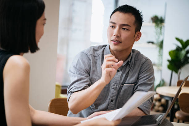 Young businessman gesturing to female coworker Asian man in his 20s talking face to face with colleague in office, explaining, discussion, collaboration asian couple talking seriously stock pictures, royalty-free photos & images