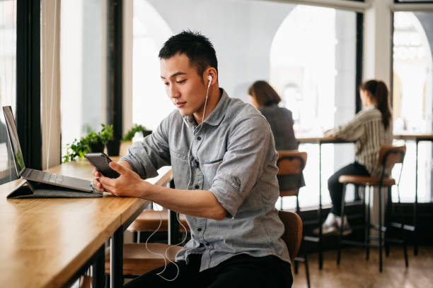 Freelance worker wearing earphones and text messaging on smartphone Professional man taking video call on mobile phone, digital nomad, connectivity, technology asian man headphones stock pictures, royalty-free photos & images
