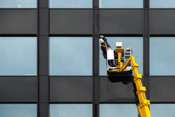 Photo of Male window cleaner cleaning glass windows on modern building high in the air on a lift platform. Worker polishing glass high in the air