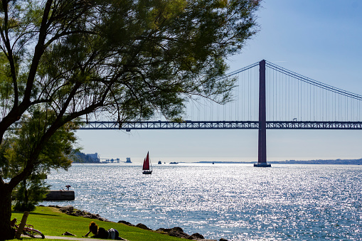 Almada, Portugal: CIRCA May 2019: Young girl lying in a park by the river in a beautiful sunny day with sailboat and suspension bridge on the background