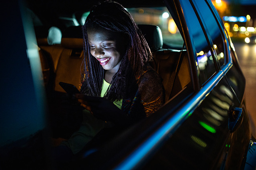 Young, beautiful, cheerful African woman driving in a taxi at night, sitting on back seat and using smart phone, smiling