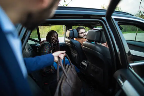 Young, handsome man entering a car, holding luggage, his friends sitting in car and waiting for him