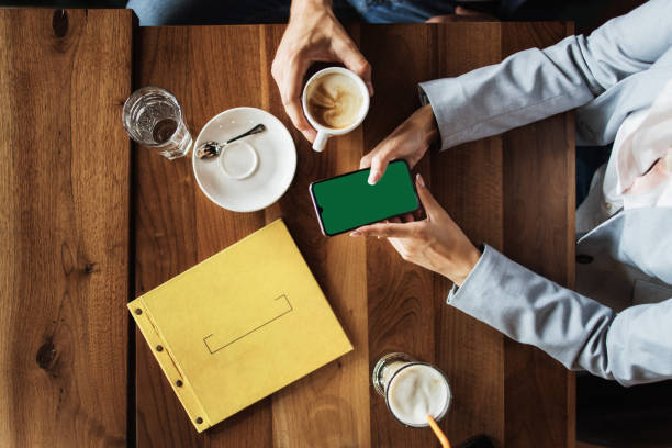 Mockup image of a business people holding smart mobile phone with blank green screen on vintage wooden table in modern cafe restaurant during meeting or lunch.