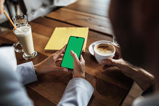 Mockup image of a business people holding smart mobile phone with blank green screen on vintage wooden table in modern cafe restaurant during meeting or lunch.