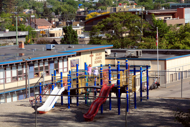Vernon,British Columbia, Canada- May 27,2019: Exterior view of school and playground of Vernon Catholic School. Small group of young children playing on outdoor playground equipment. Downtown buildings and business in background.