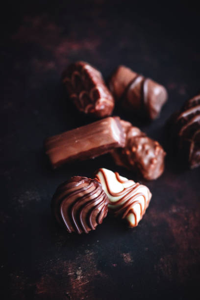 A detailed close-up of various gourmet chocolates arranged on a black plate. The image highlights the textures and shapes of dark, milk, and white chocolate pieces.