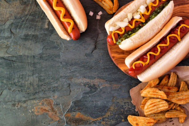 Hot dog corner border, overhead view on a dark stone background Hot dogs with toppings and potato wedges. Corner border, above view on a dark stone background with copy space. hot dog photos stock pictures, royalty-free photos & images