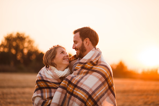 Happy man and woman are standing in field during sunset, wrapped in a blanket.