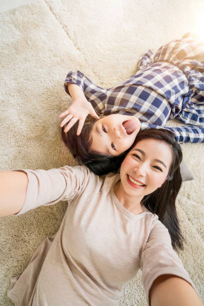 счастливая азиатская мать семьи и дочь делают фото selfie пока польза в предпосылке дома живущей комнаты. - offspring child lying on back parent стоковые фото и изображения