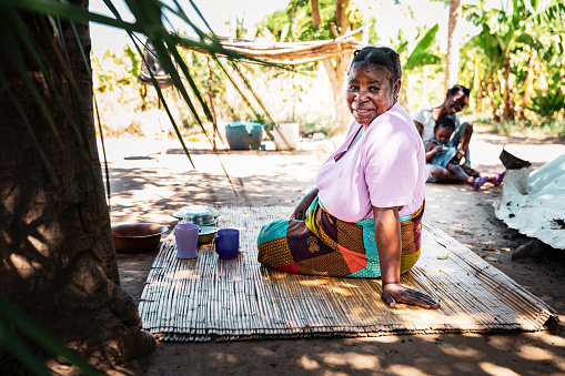 Village Life, Africa, Senior Woman - An Old Lady in a remote African village, seated on the floor mat while her daughter-in-law and granddaughter is playing in the background