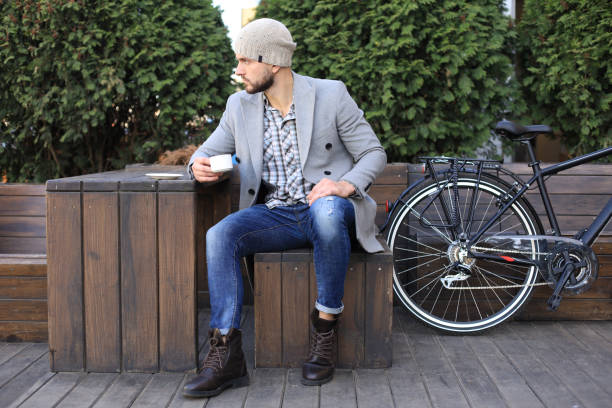 istock Handsome young man in grey coat and hat sitting on a bench relaxed drinking coffee and thinking near his bicycle. 1154548821