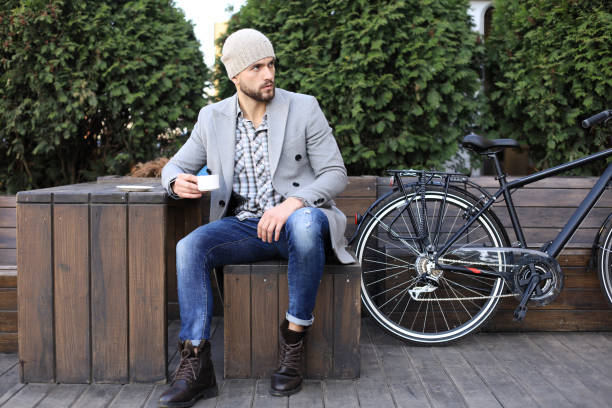 istock Handsome young man in grey coat and hat sitting on a bench relaxed drinking coffee and thinking near his bicycle. 1154549347