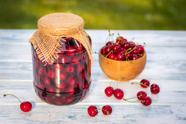 Canned cherry fruit and fresh harvested cherries in bowl on wooden table. Homemade preserved organic fruits in glass jar. Selective focus, blurry background compote photos stock pictures, royalty-free photos & images
