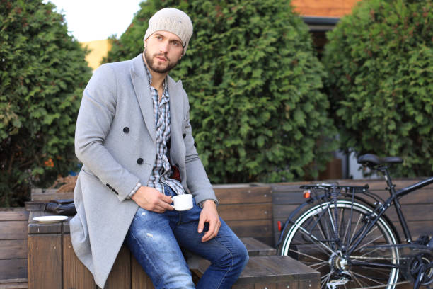 istock Handsome young man in grey coat and hat sitting on a bench relaxed drinking coffee and thinking near his bicycle. 1154550203