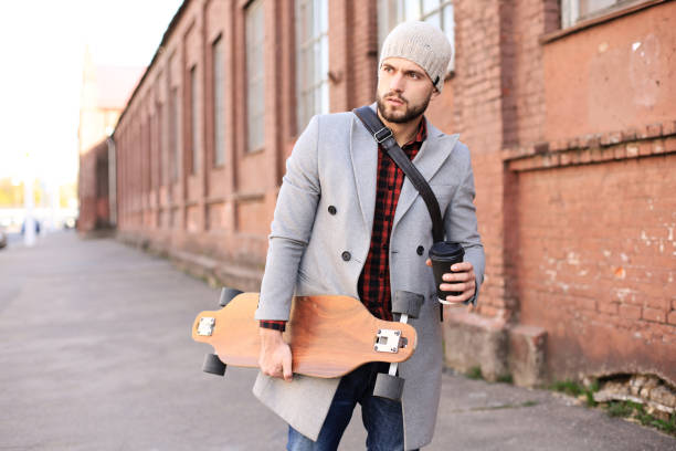 istock Handsome young man in grey coat and hat walking on the street, using longboard. 1154550463