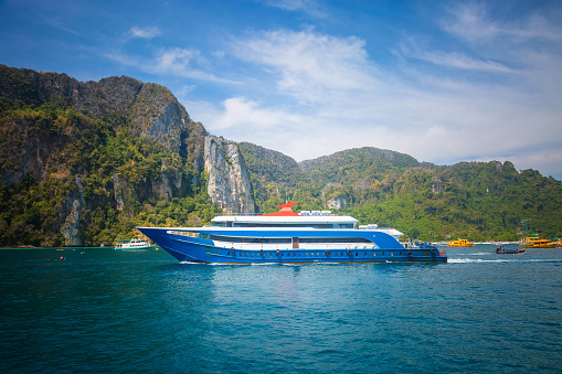 Blue and white passenger ferry ship sailing to destination point. Port with another ships and mountain on background.