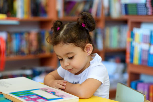 Happy child little girl reading a book. Little Girl Indoors In Front Of Books. Cute Young Toddler Sitting On A Chair Near Table and Reading Book. Child reads in a bookstore, surrounded by colorful books. Library, Shop, Shelving In Home. childhood stock pictures, royalty-free photos & images