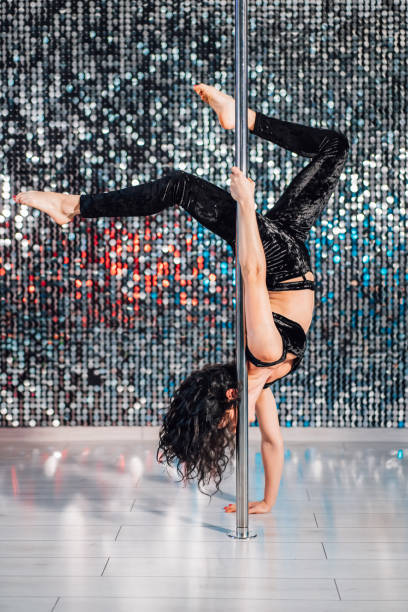 istock Woman doing tricks with pylon. Young stripper in black dances sexually with pole in studio or club. Poledance on shining wall background 1154584535