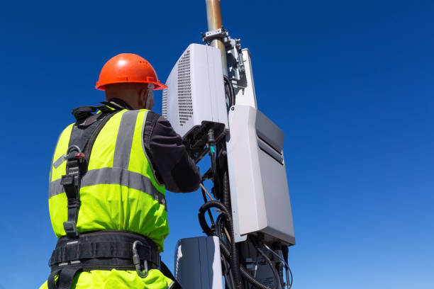 Telecommunication engineer  in helmet and uniform installs telecomunication equipment in his hand and  antennas of GSM  DCS UMTS LTE bands, outdoor radio units, optic fibers, power cables are installed on the roof. Working process of upgrading telecommuni