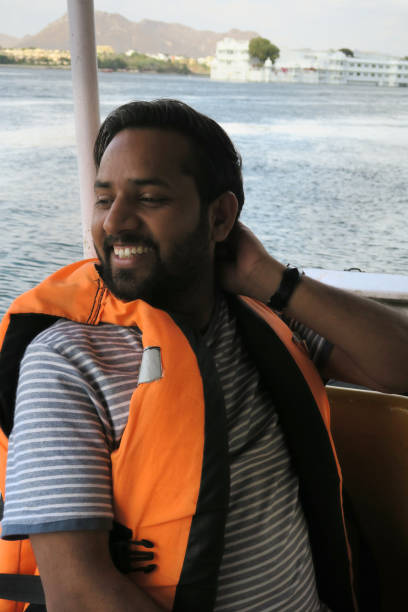 image of happy hindu indian man passenger in tourist cruise boat wearing lifejacket, orange life jacket buoyancy aid lake pichola, by taj lake palace, lake pichola udaipur, tourist boat man smiling, summer holiday vacation in udaipur, cruising lake views - motorboating sailing life jacket lake imagens e fotografias de stock