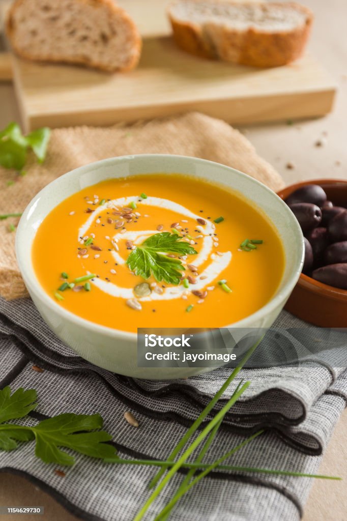 Cream of pumpkin with parsley leaves, liquid cream and seeds Pumpkin cream in a white bowl. On the table we can see some olives, a little parsley, scallion and a little bread Soup Stock Photo
