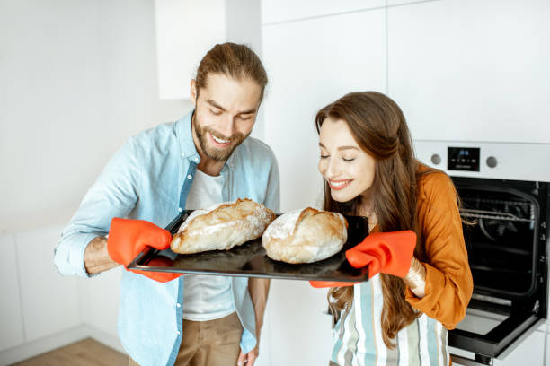 Couple with fresh breads at the kitchen Portrait of a young beautiful couple holding tray with fresh baked breads in the modern bright kitchen at home baking bread stock pictures, royalty-free photos & images
