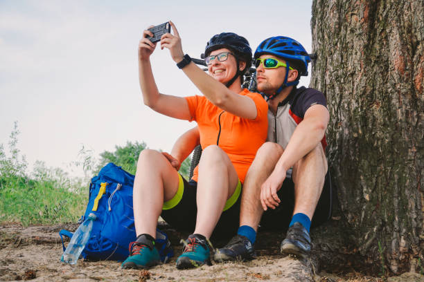 szczęśliwa para z rowerem górskim robiąc smartfon selfie na świeżym powietrzu. szczęśliwa para, która prowadzi aktywny tryb życia, zatrzymała się w lesie w pobliżu drzewa, aby zrobić zdjęcie. sport tematyczny i aktywny tryb życia - lead theme zdjęcia i obrazy z banku zdjęć