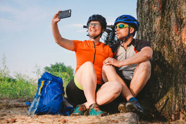 szczęśliwa para z rowerem górskim robiąc smartfon selfie na świeżym powietrzu. szczęśliwa para, która prowadzi aktywny tryb życia, zatrzymała się w lesie w pobliżu drzewa, aby zrobić zdjęcie. sport tematyczny i aktywny tryb życia - lead theme zdjęcia i obrazy z banku zdjęć