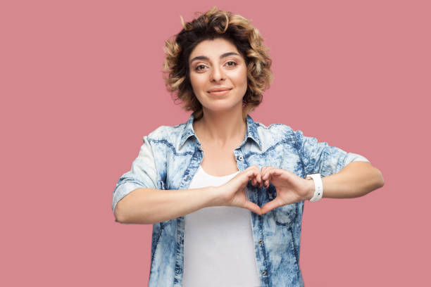 portrait de jeune femme heureuse avec la coiffure bouclée dans la chemise bleue décontractée restant avec le geste de forme de mains de coeur et regardant l’appareil-photo souriant. - eyesight concepts ideas human hand photos et images de collection