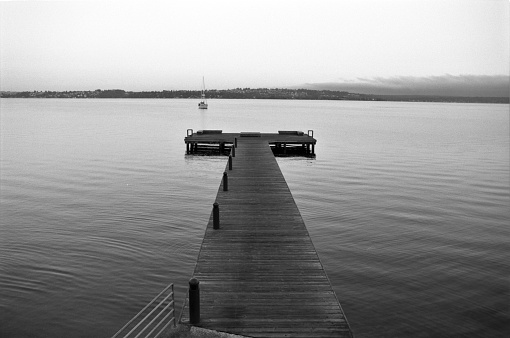Looking to north Seattle from Kirklands Brink Park Dock on Lake Washington with passing sailboat.  In black and white film.  Kirkland Washington USA.  One of many tranquil summer days in the northwest.  Serene ripples in the water make me happy.