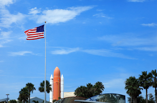 Waving American flag at Kennedy space center entrance with space rocket and palm trees over blue sky in Florida, USA