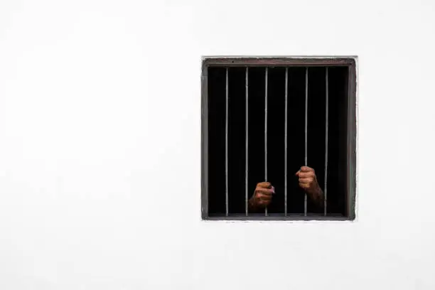 Photo of Hands of the prisoner on rusty bar of the window and prison cell white cement wall