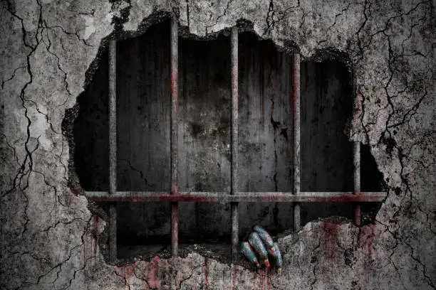 Photo of Hand of devil has stains and drops of blood and holding the damaged grungy crack and broken concrete wall in old prison metal bars, Bloody background scary