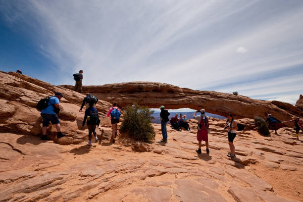 People at Mesa Arch Canyonlands National Park, Utah, USA - May 29, 2019: Mesa Arch is a pothole arch, formed when rainwater eventually broke the bonds between grains of sand and deepened surface depressions into a hole. This arch is also geologically unusual in that it sits at the top of a canyon wall. Eventually the same forces of water freezing and thawing will lead to the collapse of this arch. In this picture people are lining up to take their picture with the famous arch. Mesa Arch is on the eastern edge of the Island in the Sky Mesa. jeff goulden canyonlands national park stock pictures, royalty-free photos & images