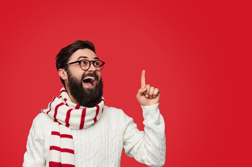 Trendy bearded man in winter clothing and scarf pointing up and looking at empty space on red background with excited face