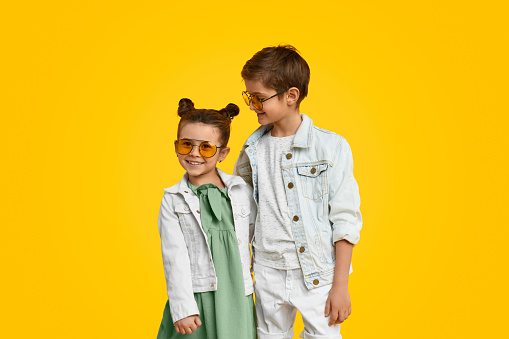 Cute boy and sweet girl wearing trendy clothes with denim jackets and sunglasses embracing on vivid yellow background