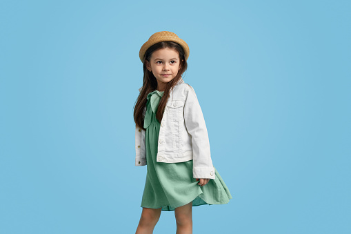 Adorable little kid in stylish dress and hat looking away while standing against blue background