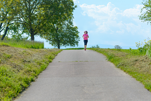 Woman working out jogging on a rural road passing through grassy fields in spring approaching the camera in the distance on the skyline