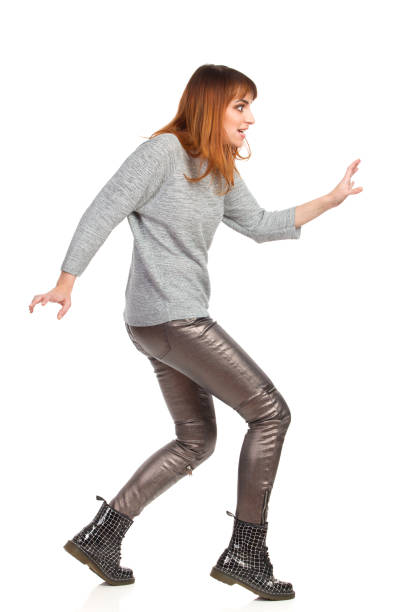 Spooky Girl Is Walking And Looking Away Spooky young woman in gray blouse, shiny pants and black boots is walking with arms outstretched and looking away. Side view. Full length studio shot isolated on white. tiptoe stock pictures, royalty-free photos & images