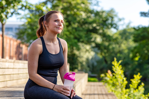 A young woman is sitting outdoors in work out clothes. She is holding a protein shake.