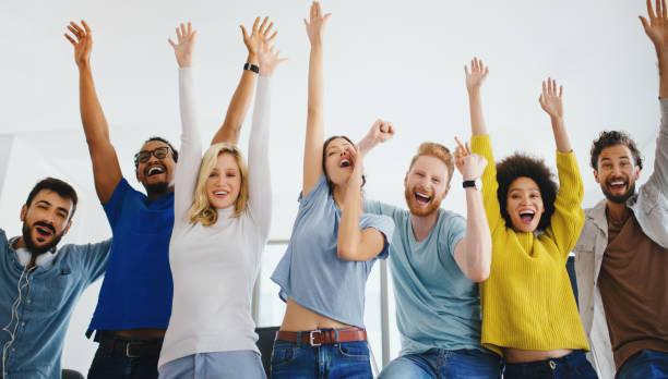 Together we achieve success. Group of cheerful young people celebrating completion of their long term project. Their are smiling and looking at camera with their arms raised. cheering group of people success looking at camera stock pictures, royalty-free photos & images
