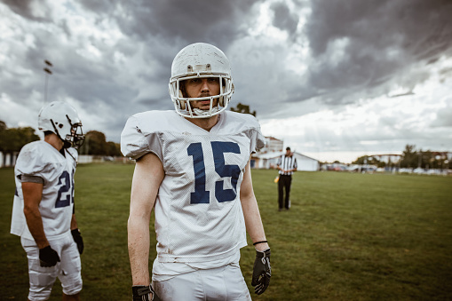 Young American football players standing on a playing field and looking at camera while his teammate is behind him.