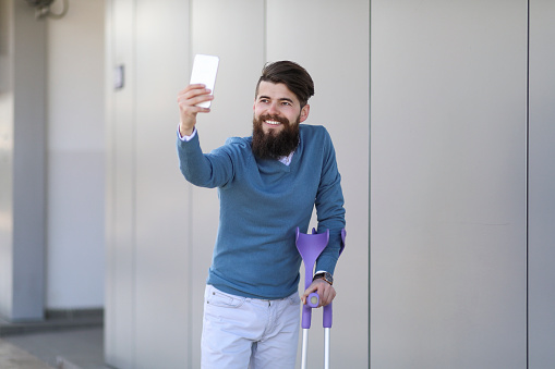 Young beautiful bearded man with walking sticks on the street, taking a stroll and enjoying sunny day. Using smartphone in outdoor environment.